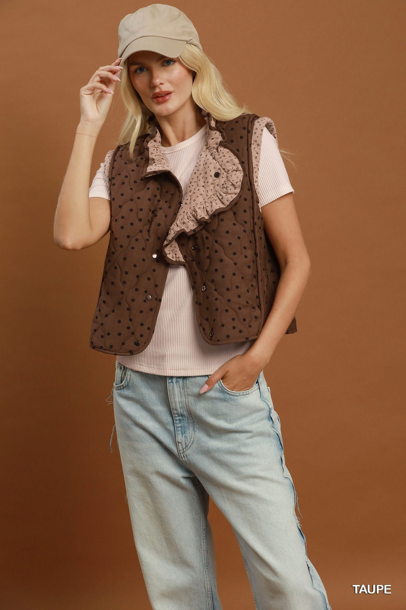 A woman models a brown sleeveless vest with a polka dot pattern and a ruffled collar, styled over a light pink t-shirt and light blue jeans against a solid brown background.