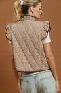 A woman shown from the back wearing a taupe quilted sleeveless vest with a ruffled high neck and small dark floral pattern, paired with a white short-sleeve top and light blue jeans against a warm brown studio background.