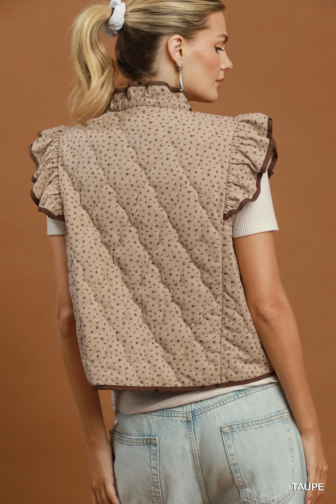A woman shown from the back wearing a taupe quilted sleeveless vest with a ruffled high neck and small dark floral pattern, paired with a white short-sleeve top and light blue jeans against a warm brown studio background.