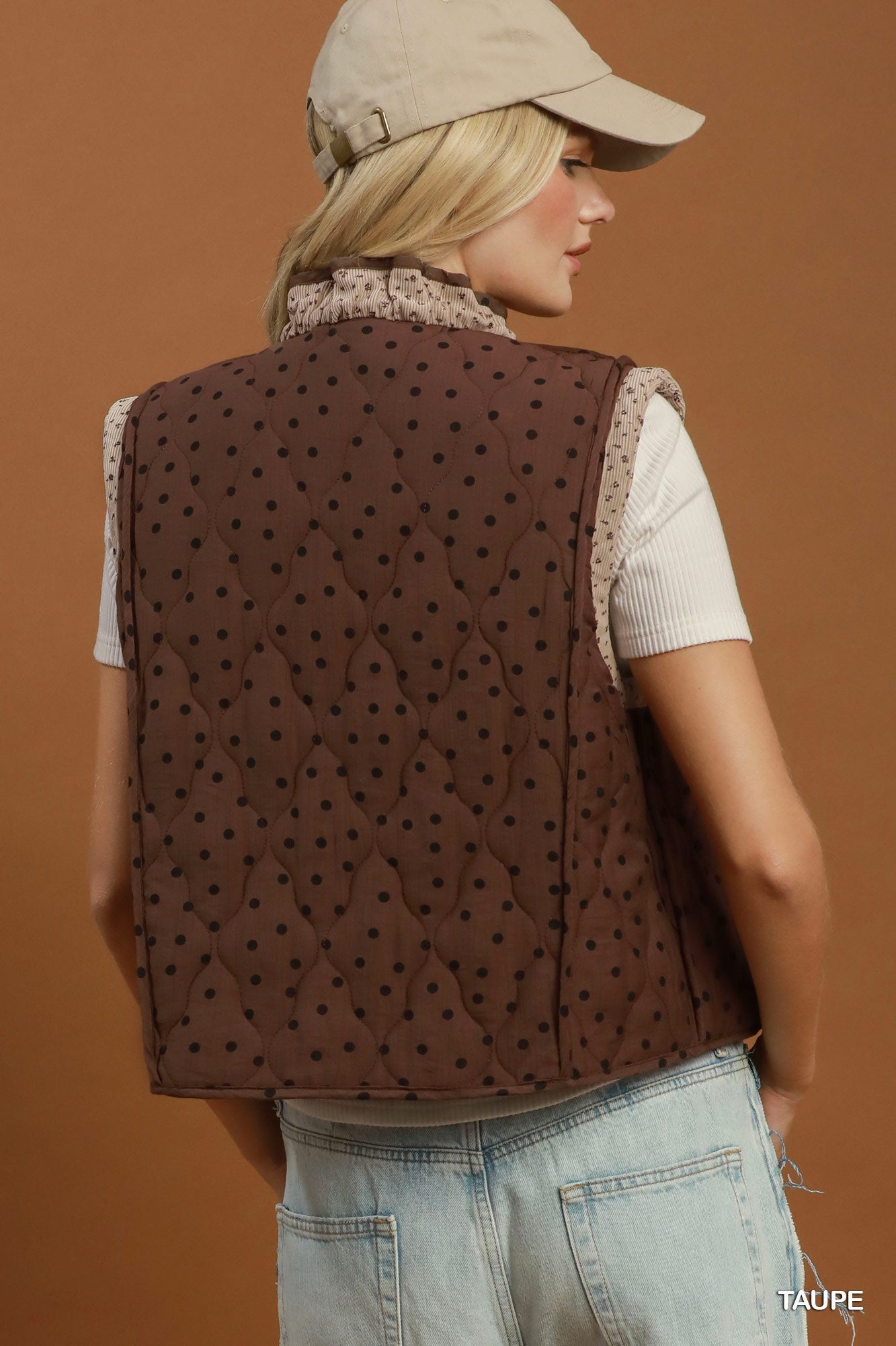 A woman shown from the back wearing a brown quilted vest with a small black polka dot pattern, layered over a white ribbed t-shirt and light blue jeans. She is also wearing a beige baseball cap.