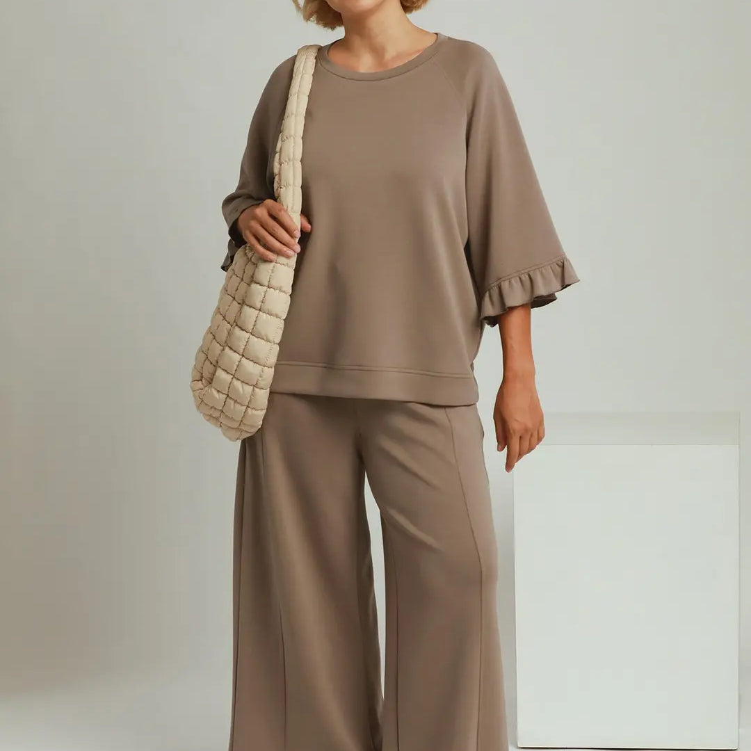 A woman modeling a two-piece lounge set in mocha brown, featuring a short-sleeve top with ruffled edges and matching wide-leg pants. She is carrying a beige quilted shoulder bag against a light gray studio background with 'PLUS SIZE' and 'MOCHA' text overlays.