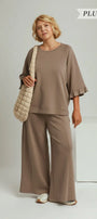 A woman modeling a two-piece lounge set in mocha brown, featuring a short-sleeve top with ruffled edges and matching wide-leg pants. She is carrying a beige quilted shoulder bag against a light gray studio background with 'PLUS SIZE' and 'MOCHA' text overlays.