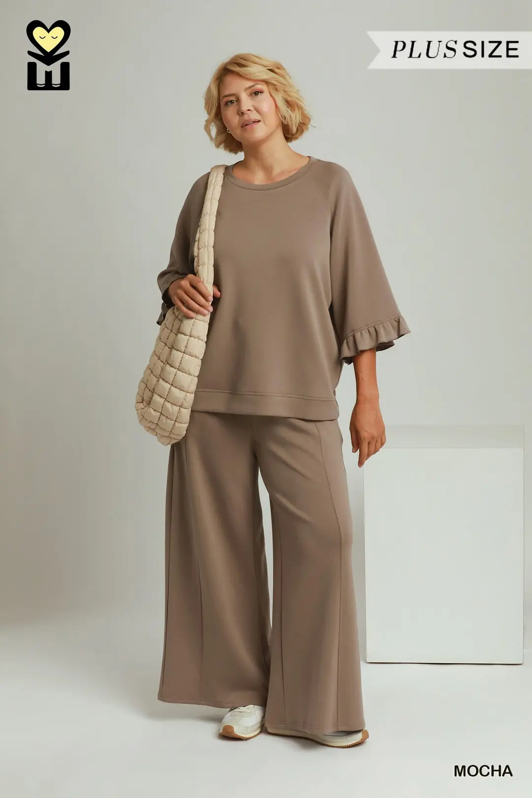 A woman modeling a two-piece lounge set in mocha brown, featuring a short-sleeve top with ruffled edges and matching wide-leg pants. She is carrying a beige quilted shoulder bag against a light gray studio background with 'PLUS SIZE' and 'MOCHA' text overlays.