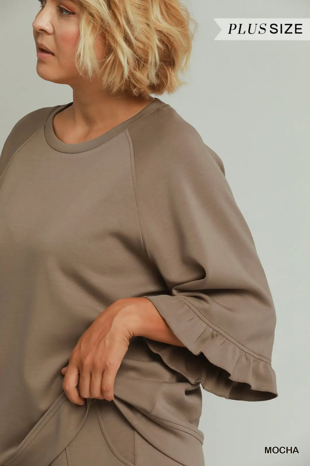 A woman wearing a loose, oversized taupe-brown top with wide three-quarter sleeves and ruffled cuffs, shown from the side against a light gray studio background. Text overlays read 'PLUSSIZE' and 'MOCHA'.