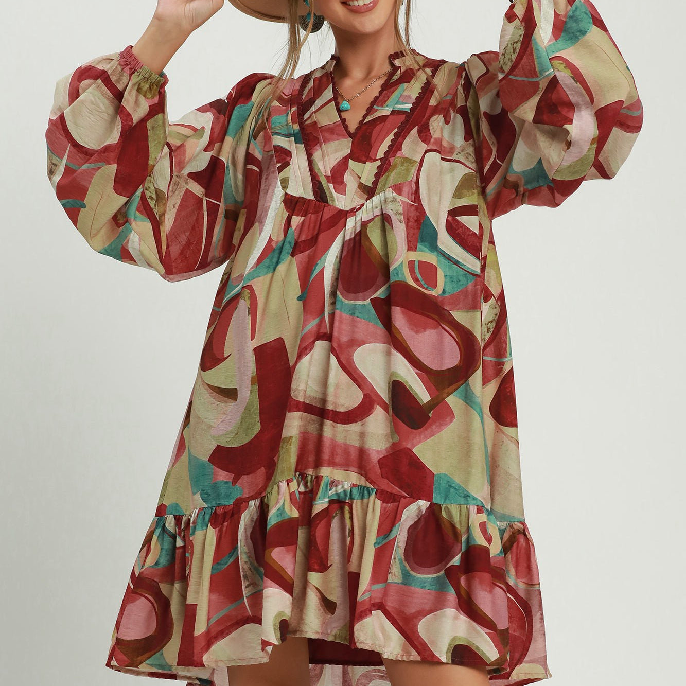 A woman modeling a bohemian-style long-sleeve dress with an abstract multicolor print in red, beige, pink, and teal, wearing a wide-brimmed tan hat against a light gray studio background.