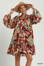 A woman modeling a bohemian-style long-sleeve dress with an abstract multicolor print in red, beige, pink, and teal, wearing a wide-brimmed tan hat against a light gray studio background.