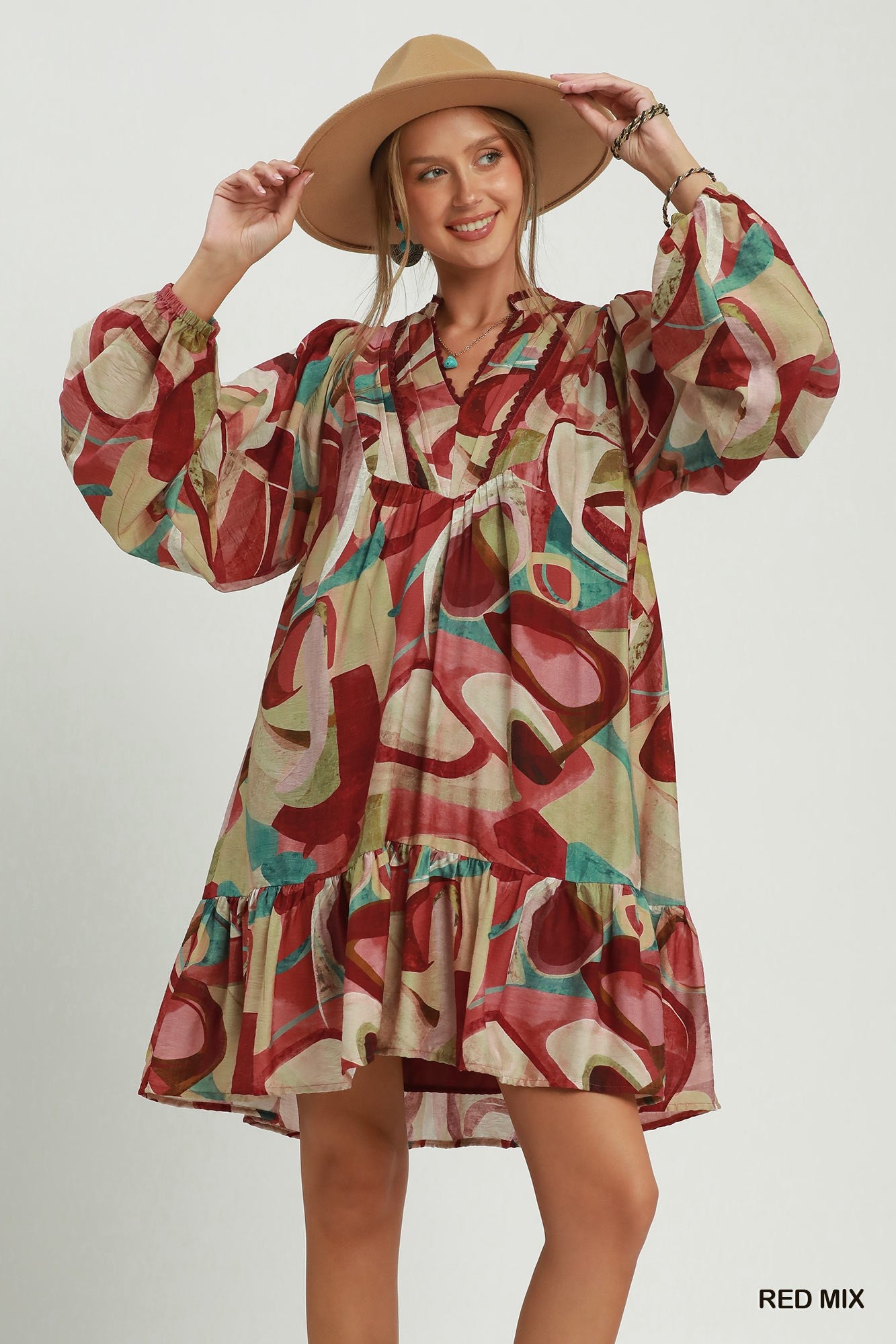 A woman modeling a bohemian-style long-sleeve dress with an abstract multicolor print in red, beige, pink, and teal, wearing a wide-brimmed tan hat against a light gray studio background.