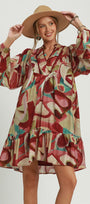 A woman modeling a bohemian-style long-sleeve dress with an abstract multicolor print in red, beige, pink, and teal, wearing a wide-brimmed tan hat against a light gray studio background.