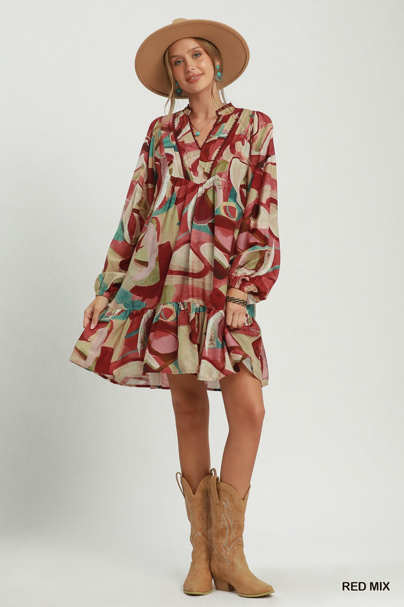 A woman modeling a long-sleeve, collared mini dress with a multicolor abstract print in red, beige, and teal, styled with a tan cowboy hat and tan cowboy boots against a light gray studio background.