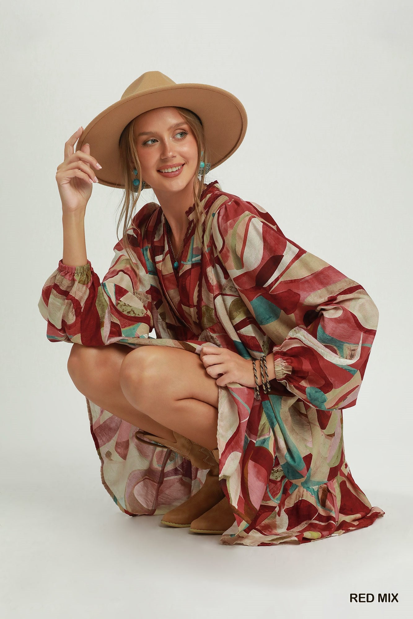 A woman crouches in a studio wearing a loose, long-sleeve bohemian-style dress with an abstract red, beige, and teal print, along with a wide-brim tan hat. The text 'RED MIX' is printed in the bottom right corner.