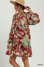 A woman wearing a long-sleeve bohemian mini dress with an abstract multicolor print in red, beige, and teal, paired with a tan wide-brim hat against a light gray studio background.
