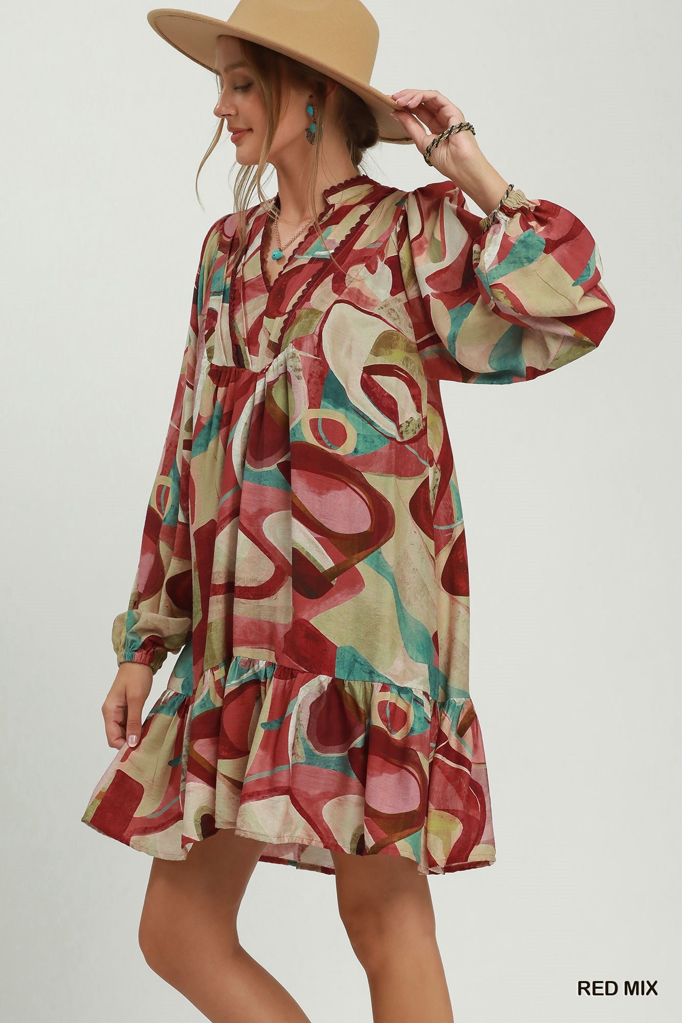 A woman wearing a long-sleeve bohemian mini dress with an abstract multicolor print in red, beige, and teal, paired with a tan wide-brim hat against a light gray studio background.