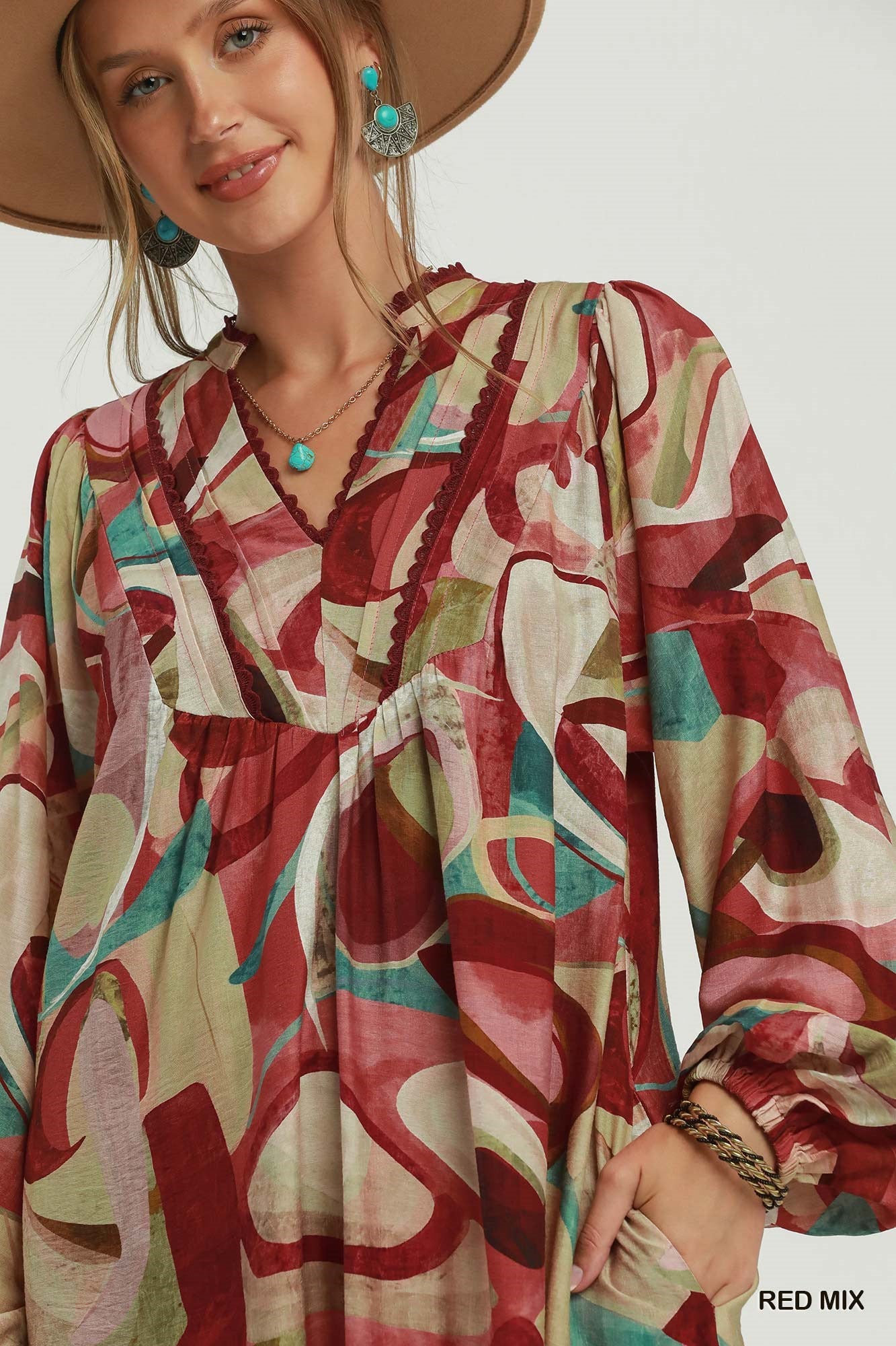 A woman wearing a loose, long-sleeve bohemian-style dress with an abstract watercolor print in red, beige, and teal, paired with a wide-brimmed tan hat and turquoise jewelry. A 'RED MIX' label is visible in the bottom right corner.