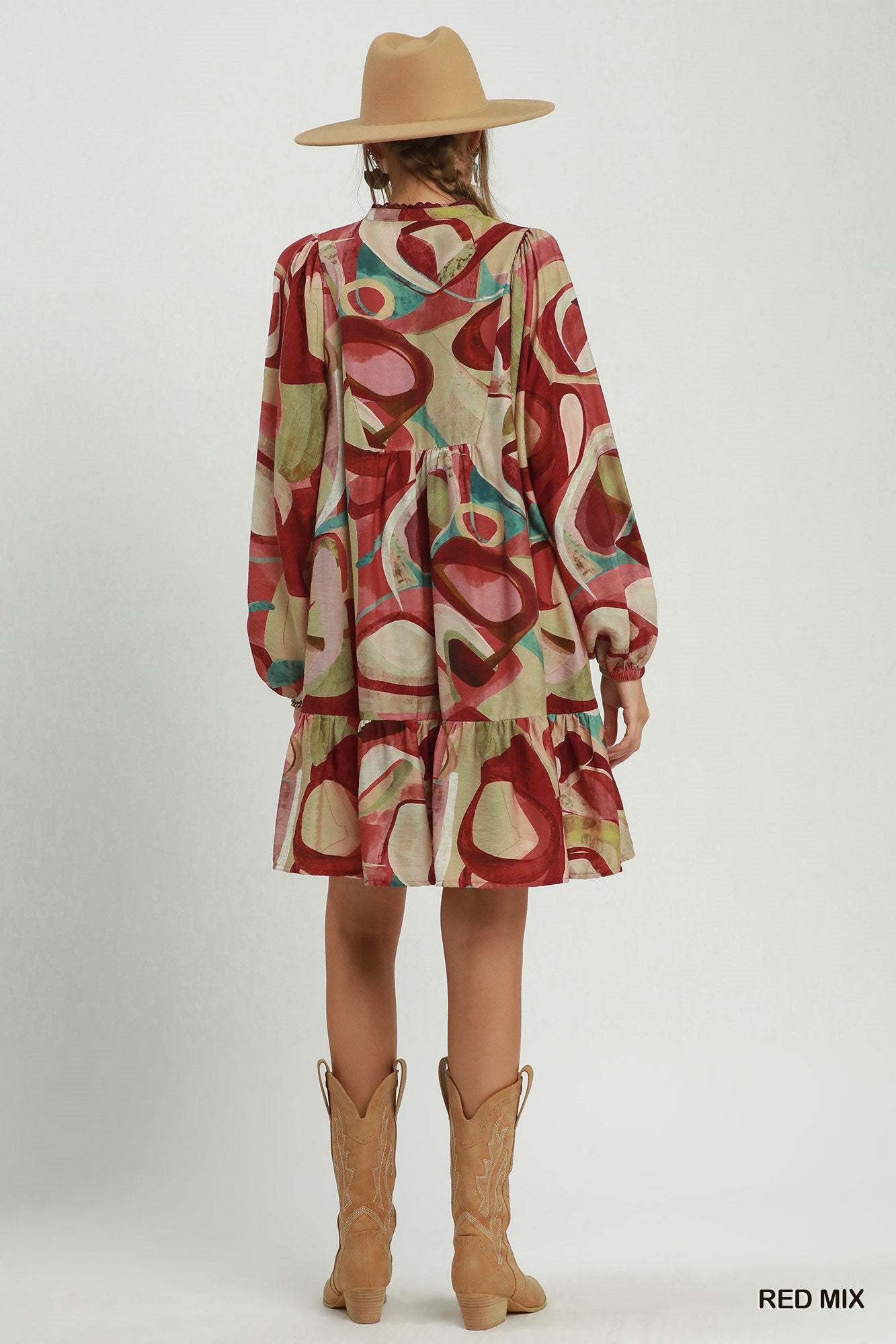 A model shown from the back wearing a tiered long-sleeve dress with an abstract red, beige, and teal pattern, styled with a tan hat and tan cowboy boots against a light gray studio background.