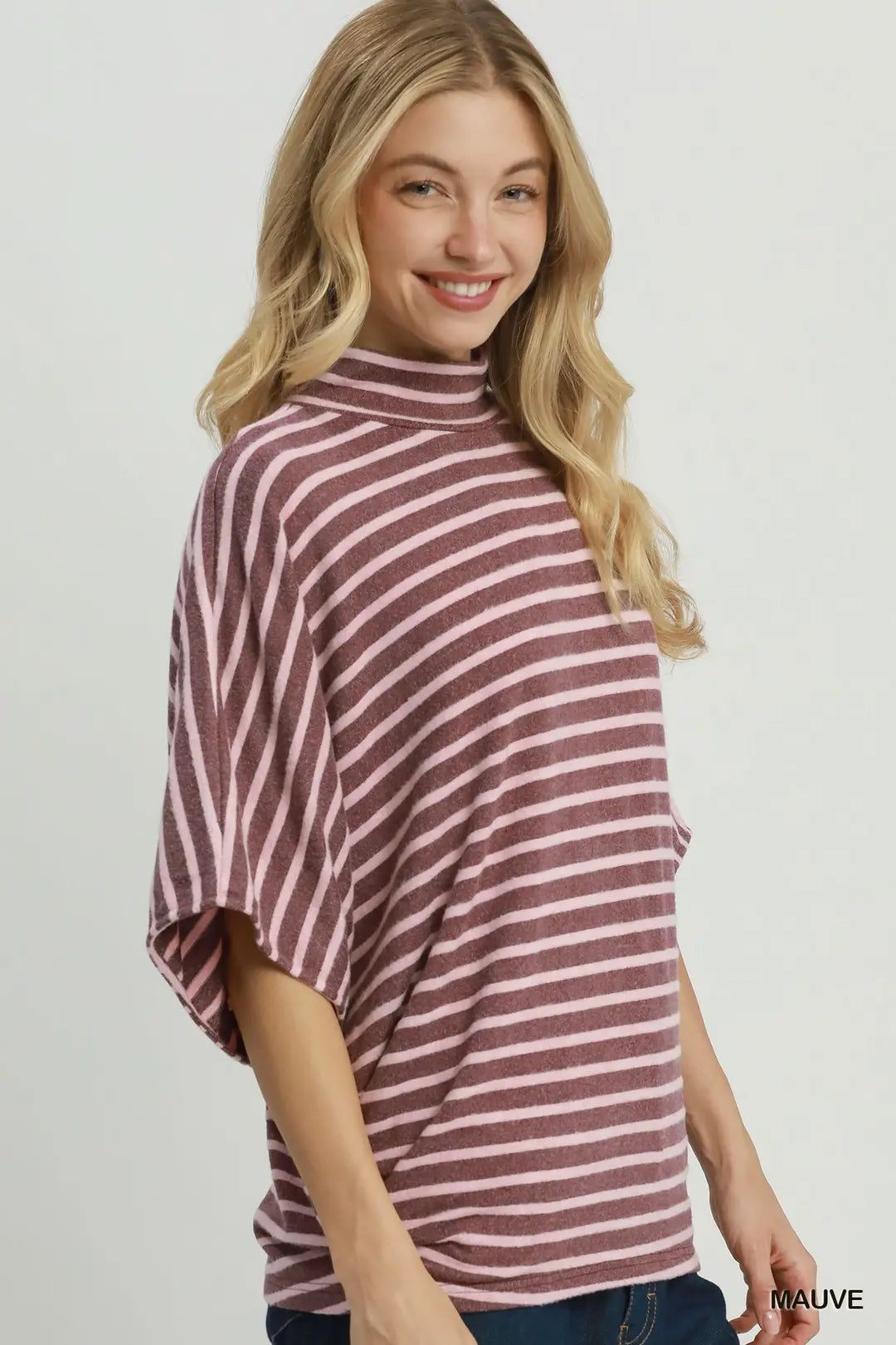 A woman wearing a mauve and white striped turtleneck top with wide three-quarter sleeves, paired with dark blue jeans, against a light gray studio background.