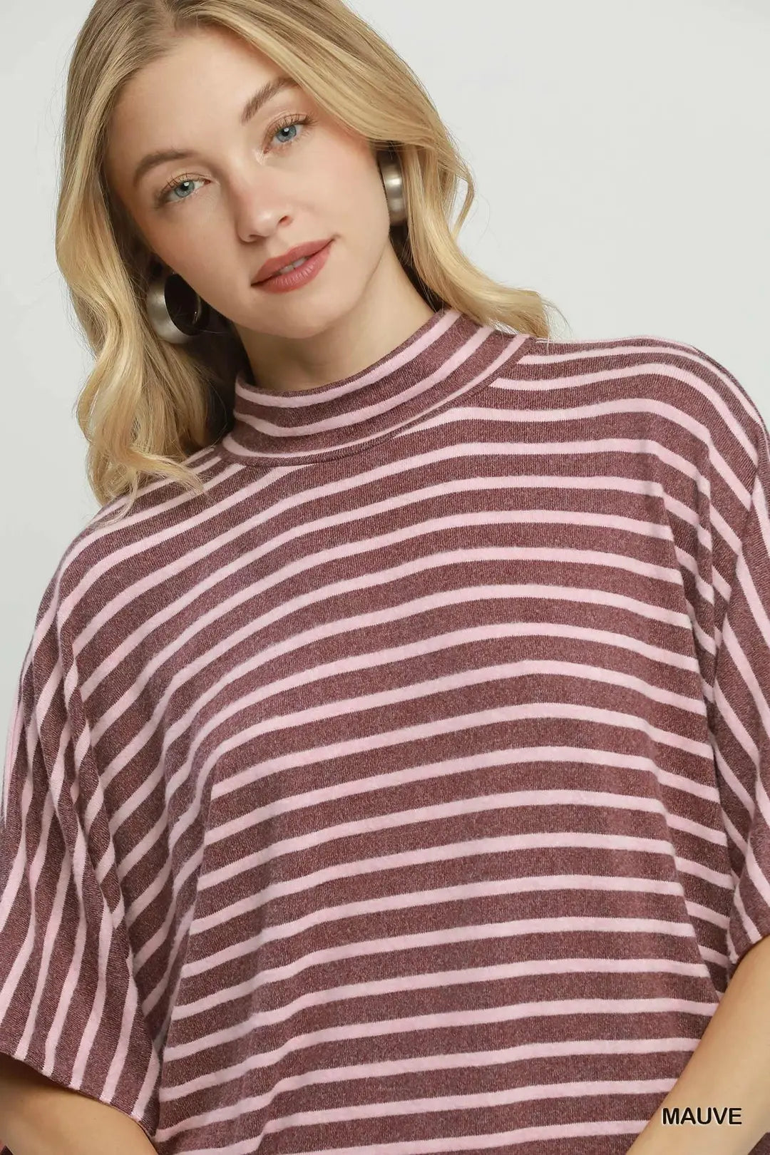 A woman wearing a loose-fitting short-sleeve mock neck top with horizontal stripes in dusty mauve and light pink, shown against a plain light gray background.