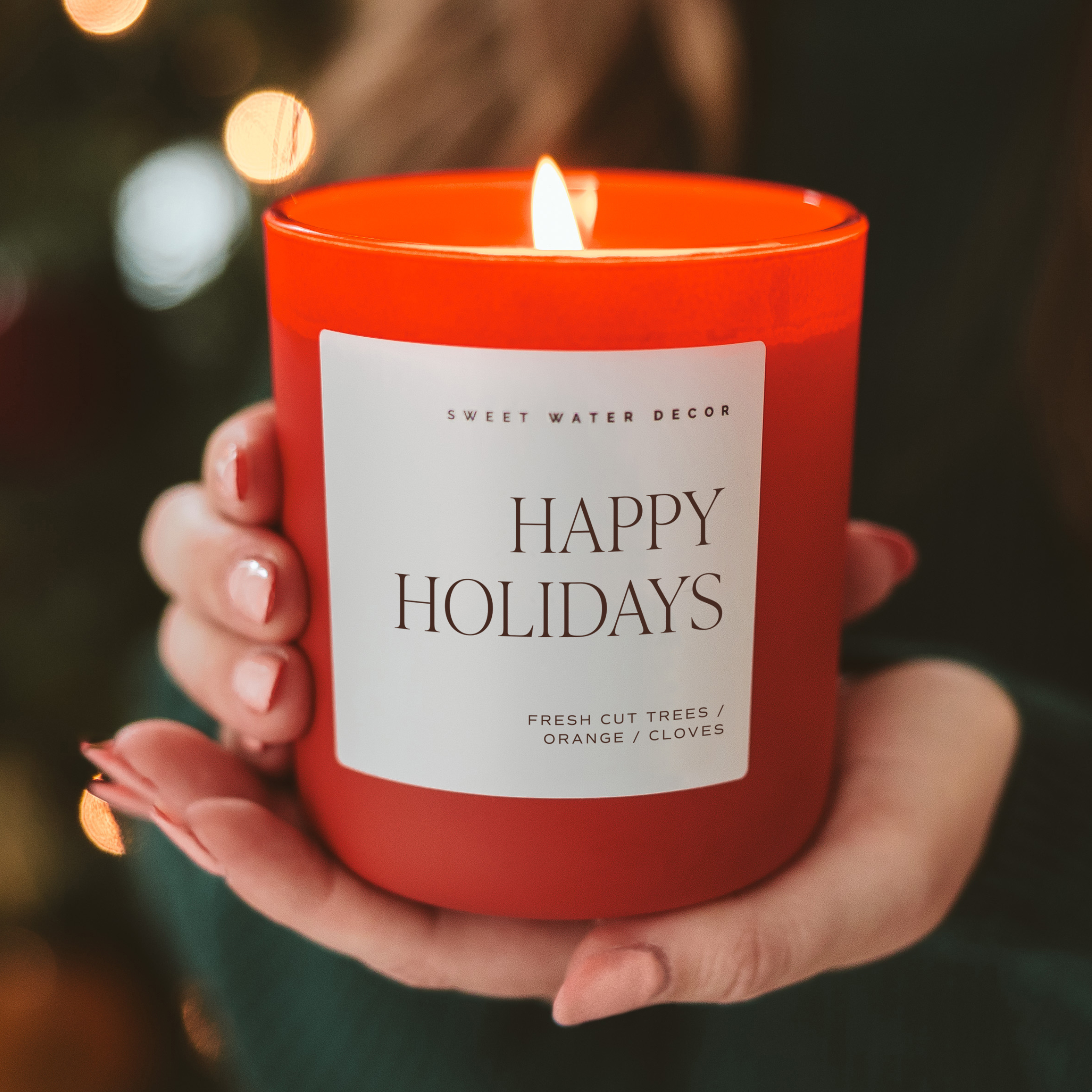 Red candle labeled 'Happy Holidays' held by a hand with blurred lights in the background