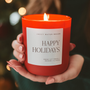 Red candle labeled 'Happy Holidays' held by a hand with blurred lights in the background