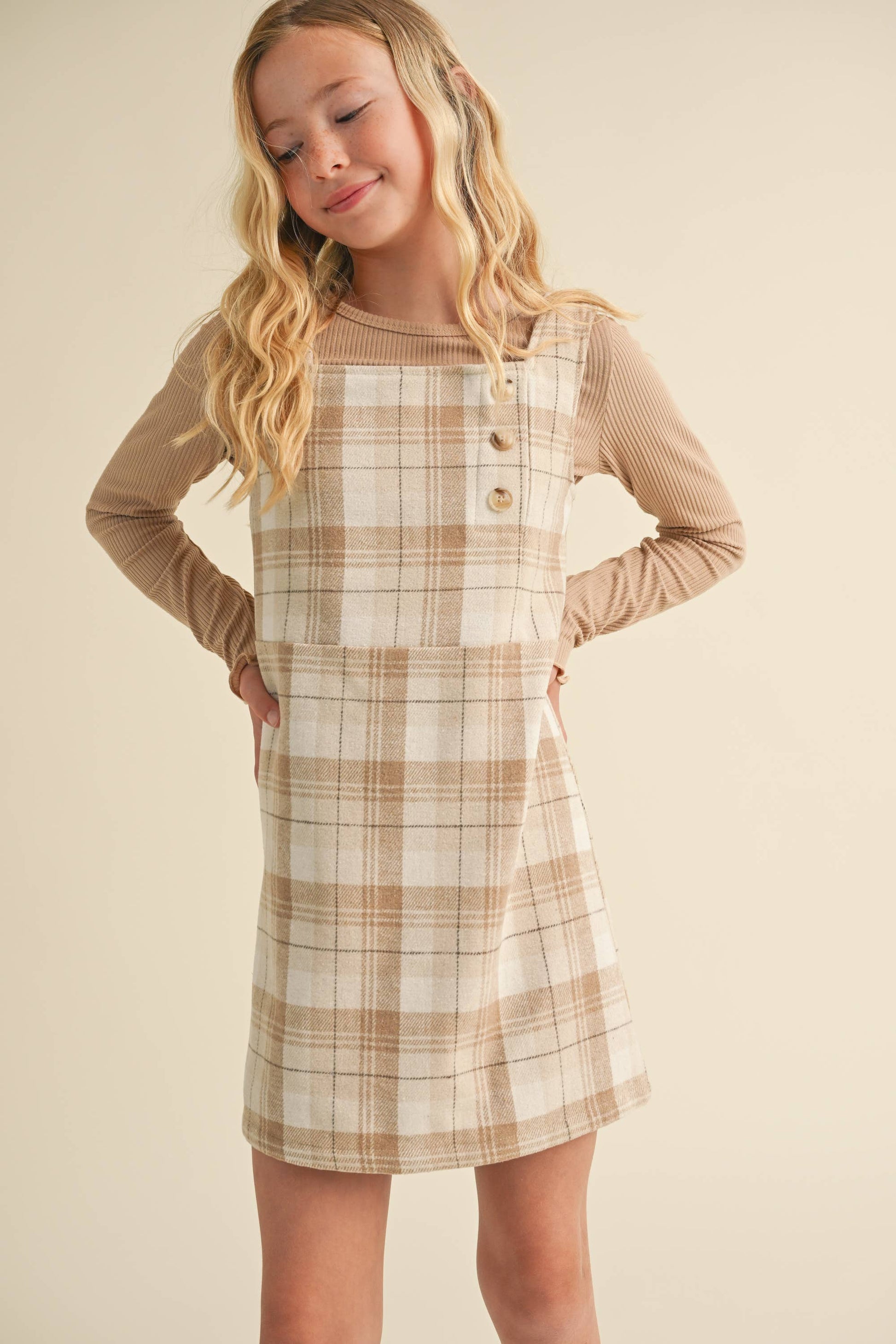 Young girl wearing a plaid dress over a long-sleeve shirt against a beige background