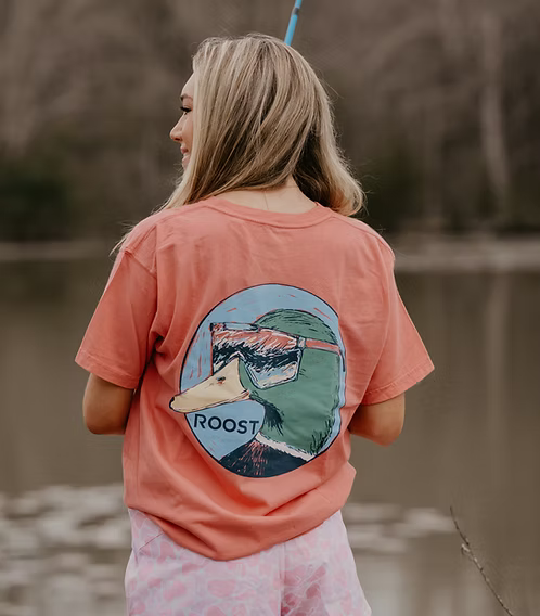 Woman wearing a pink t-shirt with a duck graphic and 'Roost' text, standing by a body of water.