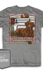 A gray short-sleeve graphic t-shirt featuring a large front print of a brown dog, a football, and the text 'FIELDSTONE'. A matching gray fabric pocket patch with the Fieldstone logo is shown in the bottom-left corner against a white background.