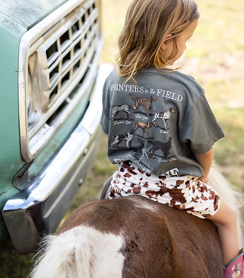 Child sitting on a horse wearing a shirt with 'Painters in the Field' design, standing next to a vintage truck.