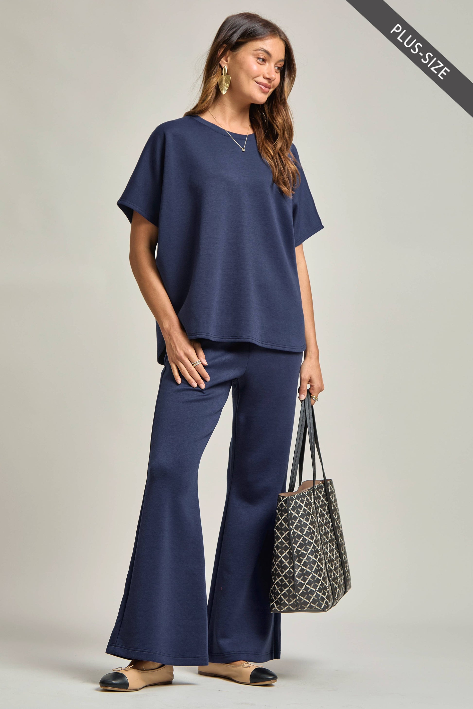 Woman wearing a navy outfit with wide-leg pants and holding a patterned bag on a plain background