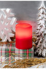 A red LED flameless pillar candle sits on a plaid gift box surrounded by Christmas decorations, including a white snowflake, a small snowy tree, and gold tinsel, against a gray background.