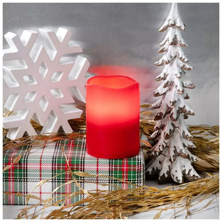 A red LED flameless pillar candle sits on a plaid gift box surrounded by Christmas decorations, including a white snowflake, a small snowy tree, and gold tinsel, against a gray background.