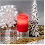 A red LED flameless pillar candle sits on a plaid gift box surrounded by Christmas decorations, including a white snowflake, a small snowy tree, and gold tinsel, against a gray background.