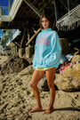 Person wearing a light blue sweatshirt with pink text on a beach.