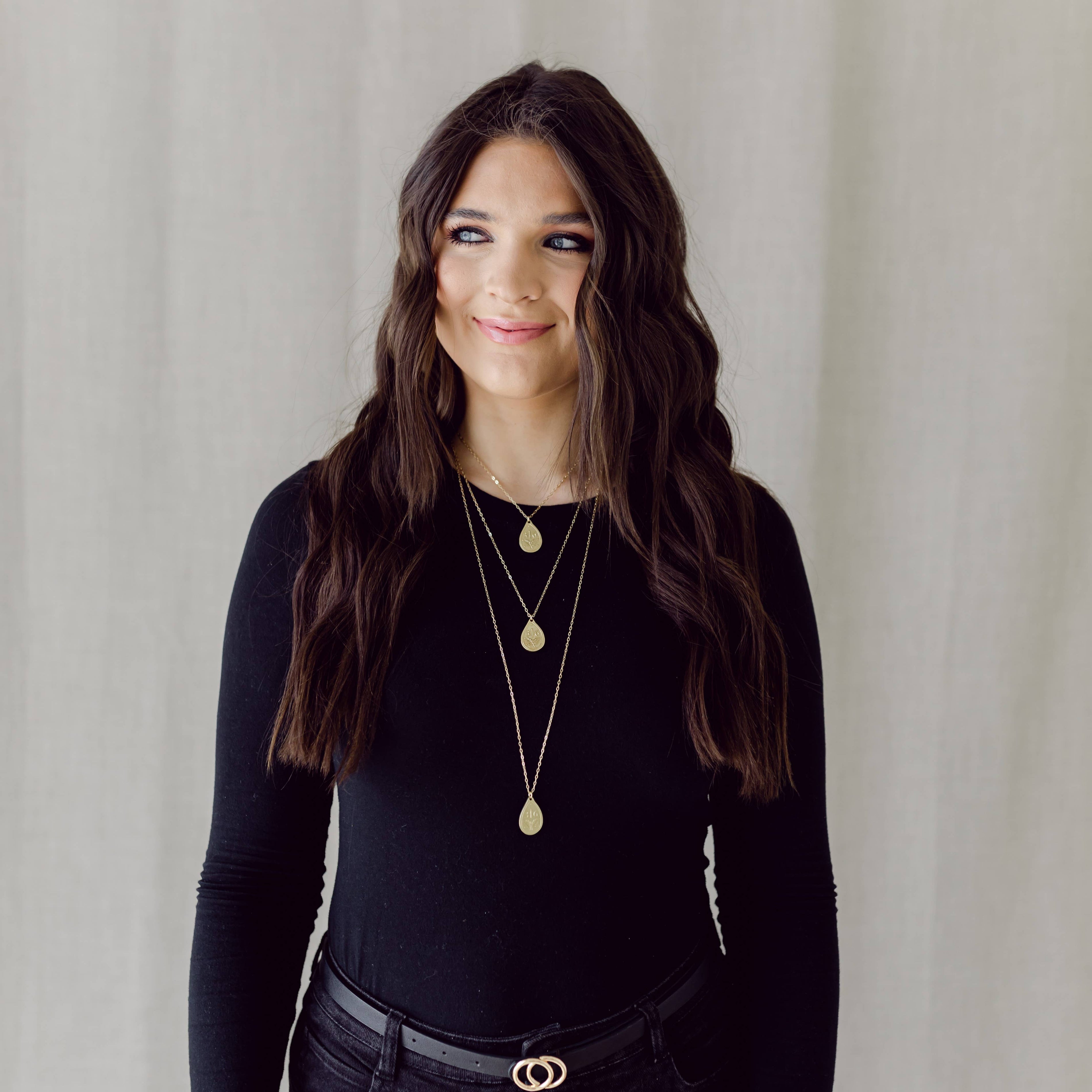 Woman wearing a black long-sleeve top and gold necklaces against a neutral background