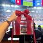 A person holds a clear tote bag with a large red ribbon bow and red straps, with a blurred stadium background visible behind it.