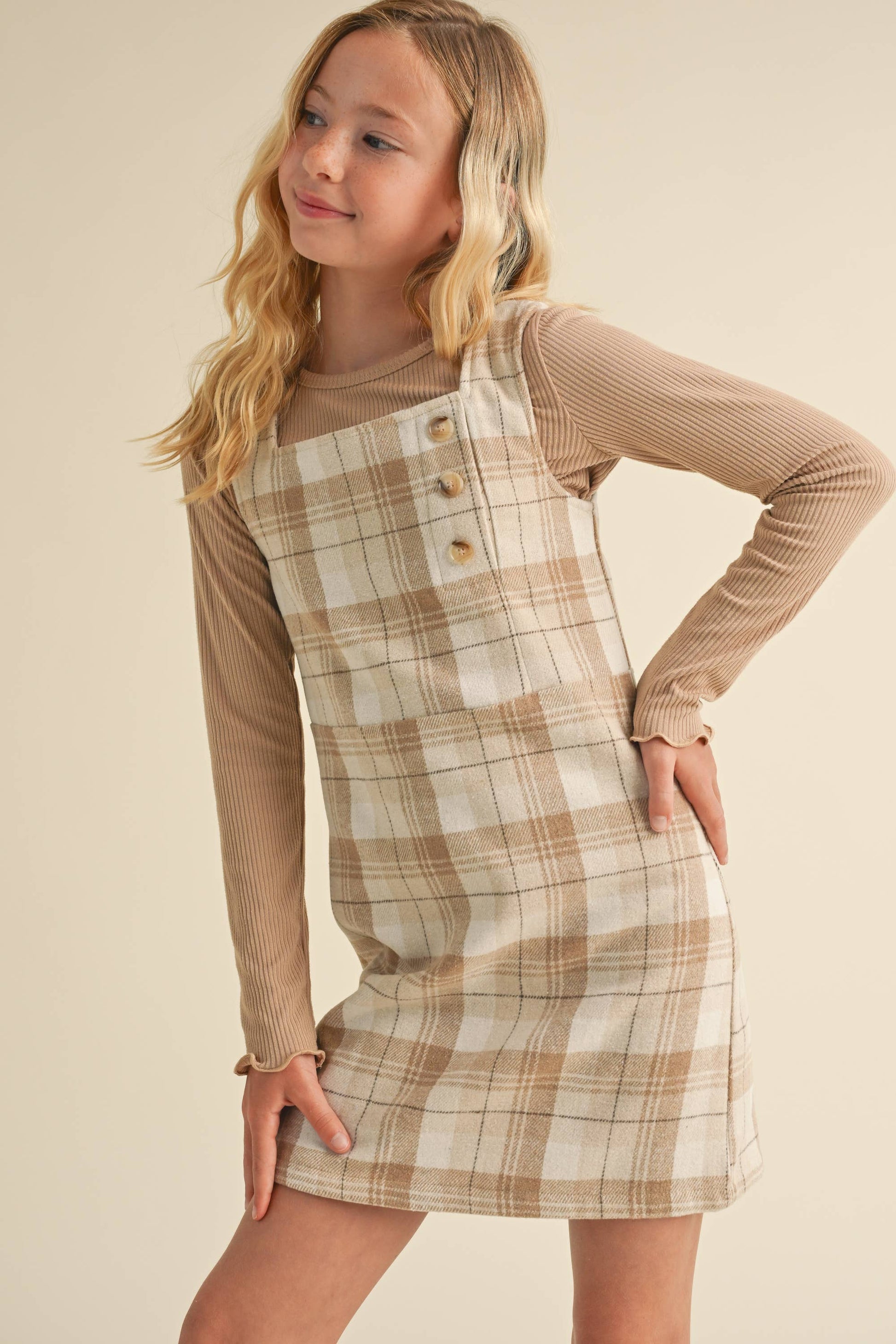 Young girl wearing a plaid dress over a long-sleeve shirt against a beige background