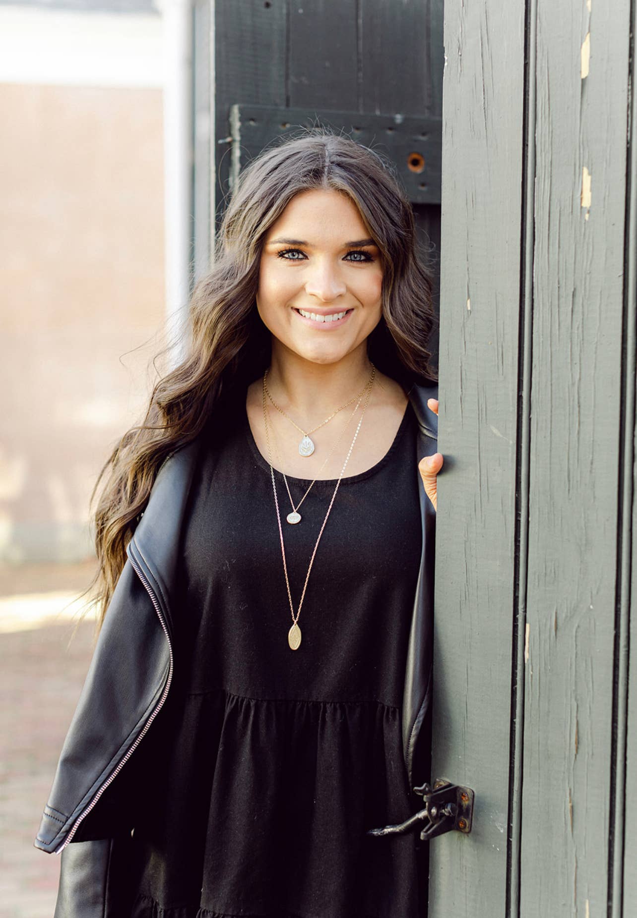 Woman wearing a black top and layered necklaces, standing next to a wooden door.
