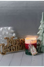 A holiday tabletop arrangement featuring a gold 'Merry Christmas' sign, a white snowflake decoration, a lit red and green candle with a hanging ornament, and a mint green ceramic Christmas tree on a light gray background.