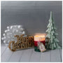 A holiday tabletop arrangement featuring a gold 'Merry Christmas' sign, a white snowflake decoration, a lit red and green candle with a hanging ornament, and a mint green ceramic Christmas tree on a light gray background.