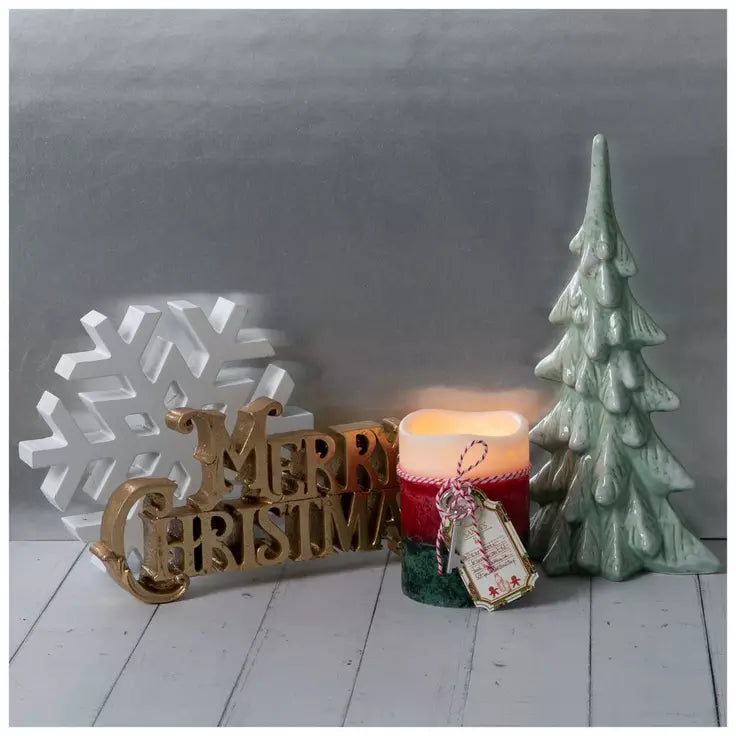 A holiday tabletop arrangement featuring a gold 'Merry Christmas' sign, a white snowflake decoration, a lit red and green candle with a hanging ornament, and a mint green ceramic Christmas tree on a light gray background.