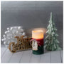 A lit Christmas-themed candle with a red and dark green body sits on a white wooden surface, styled with a gold 'Merry Christmas' sign, a white snowflake ornament, and a green tree decoration against a gray backdrop.