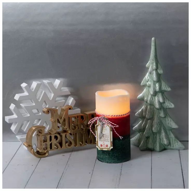 A lit Christmas-themed candle with a red and dark green body sits on a white wooden surface, styled with a gold 'Merry Christmas' sign, a white snowflake ornament, and a green tree decoration against a gray backdrop.