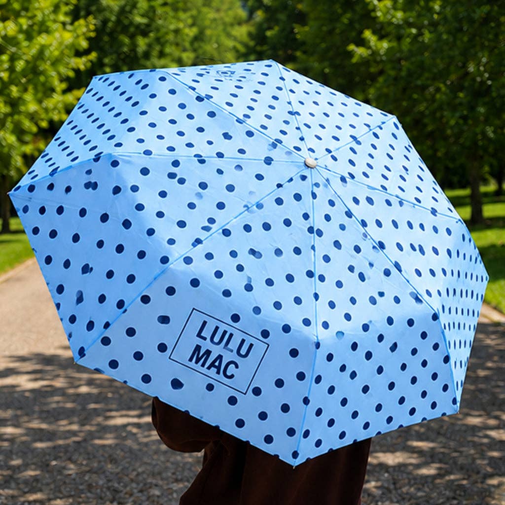 Blue polka dot umbrella with 'LULU MAC' branding held outdoors.