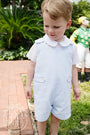 Young boy in a light blue romper standing outdoors with greenery in the background
