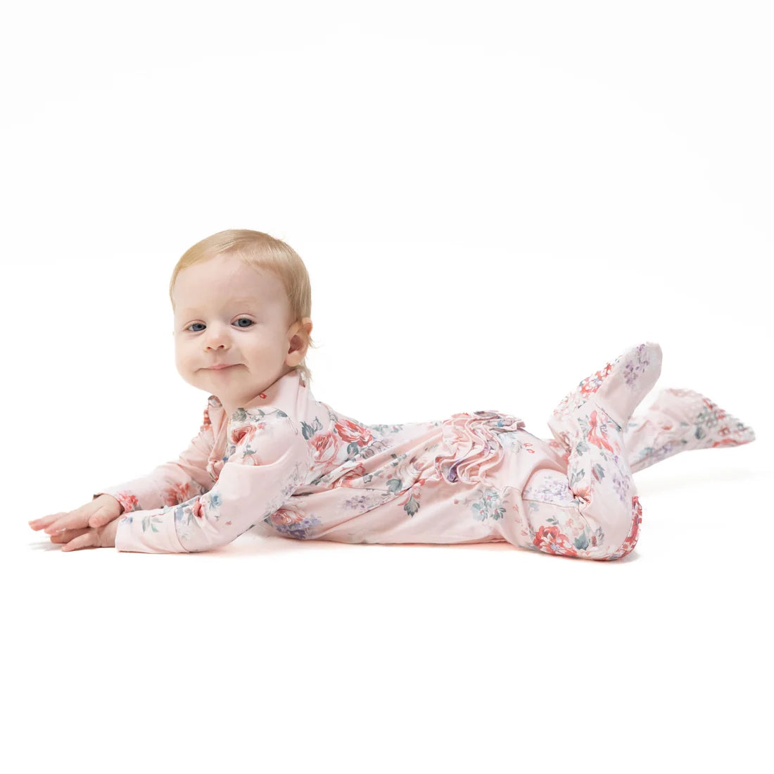 A baby lying on their stomach wearing a light pink footed sleeper with a delicate floral pattern, photographed against a seamless white background.