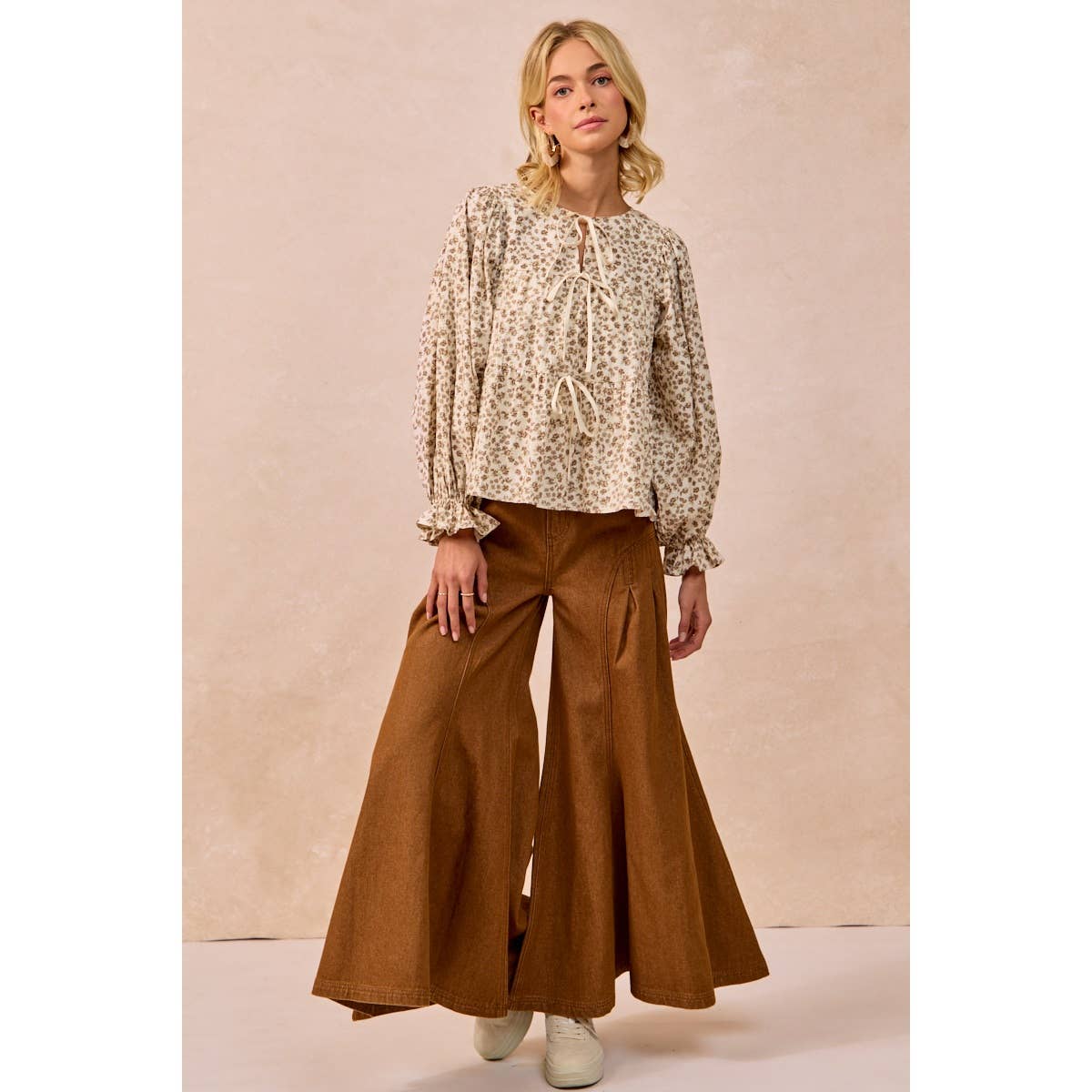 A woman modeling a beige and brown floral print long-sleeve blouse with a tie-neck detail, paired with wide-leg brown pants, against a textured beige studio backdrop.