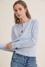 A woman wearing a light blue knit cardigan sweater with button details and blue jeans, posed against a plain off-white background.