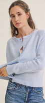 A woman wearing a light blue knit cardigan sweater with button details and blue jeans, posed against a plain off-white background.
