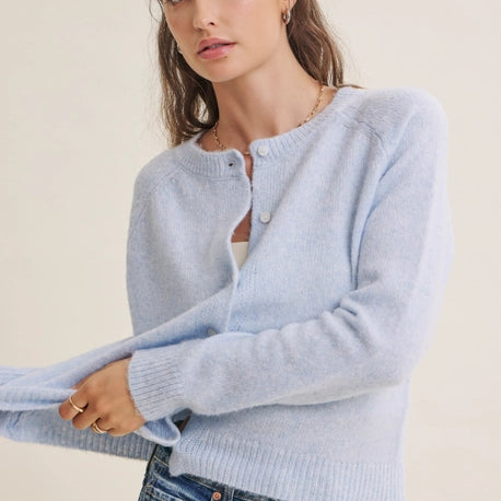 A woman wearing a light blue knit cardigan sweater with button details and blue jeans, posed against a plain off-white background.
