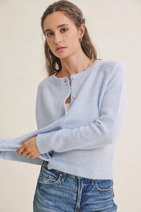 A woman wearing a light blue knit cardigan sweater with button details and blue jeans, posed against a plain off-white background.