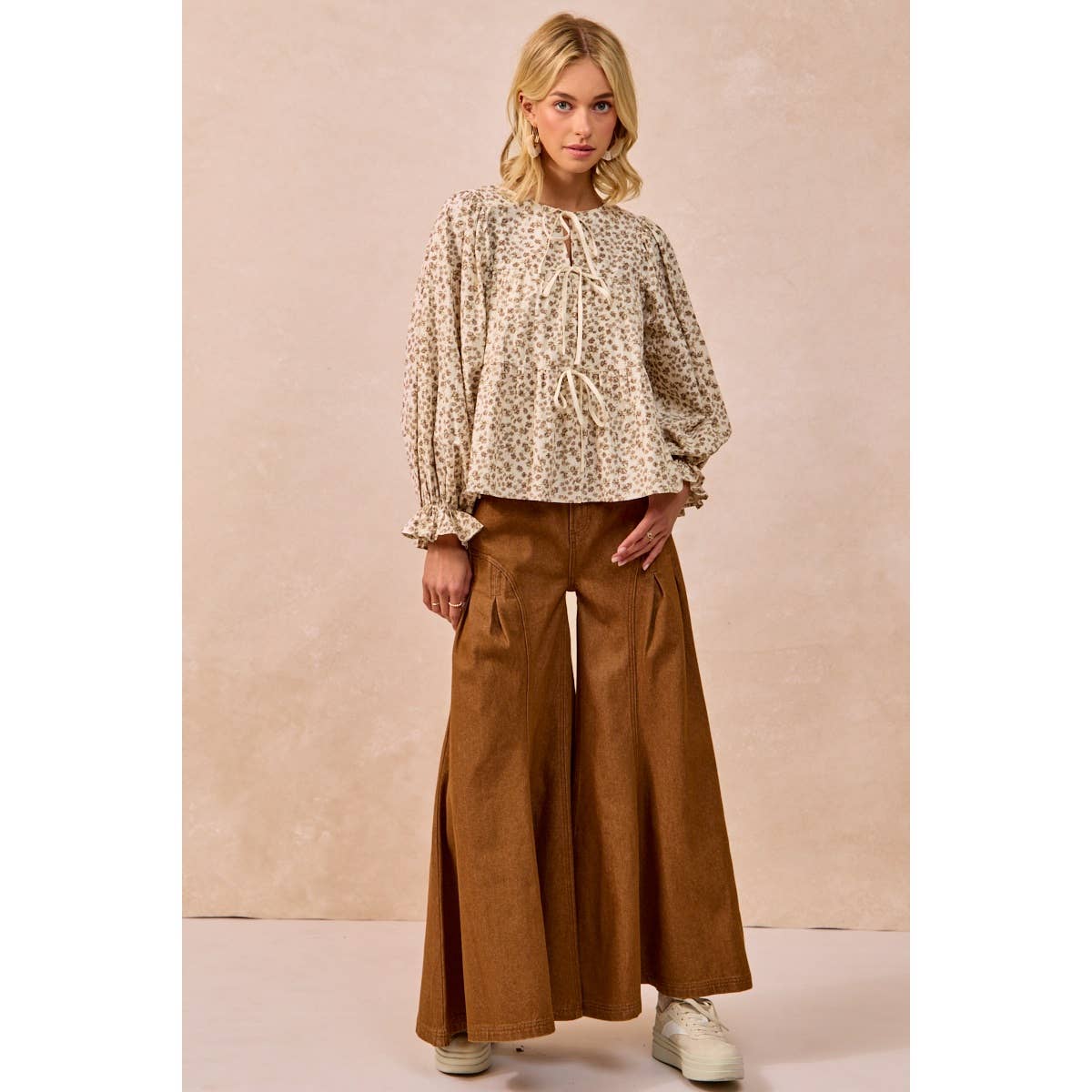 A woman modeling a beige and brown floral long-sleeve blouse with a tie neckline, paired with wide-leg brown pants, standing against a textured beige studio backdrop.