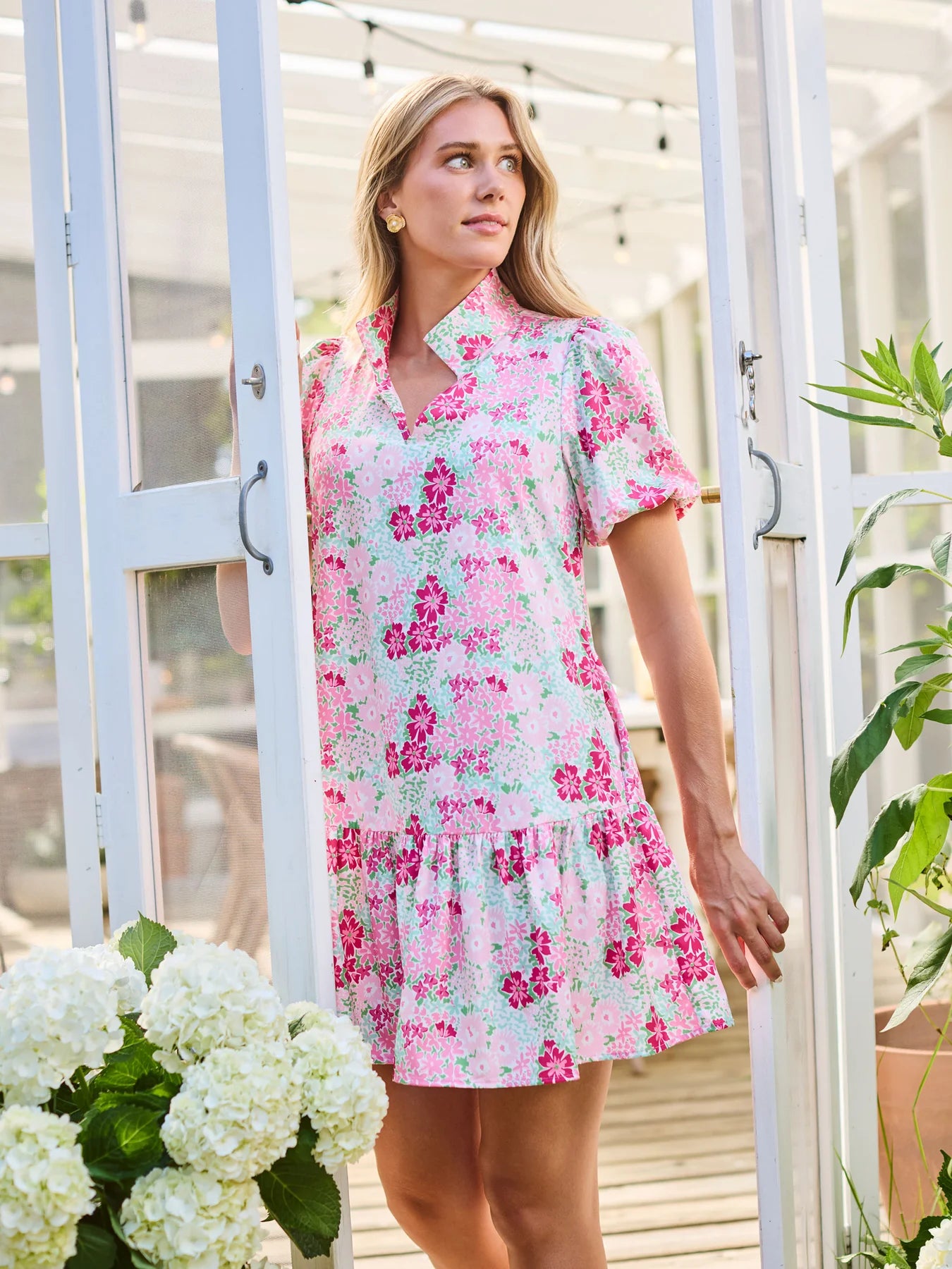 Woman wearing a floral dress standing in a greenhouse with plants and flowers around.
